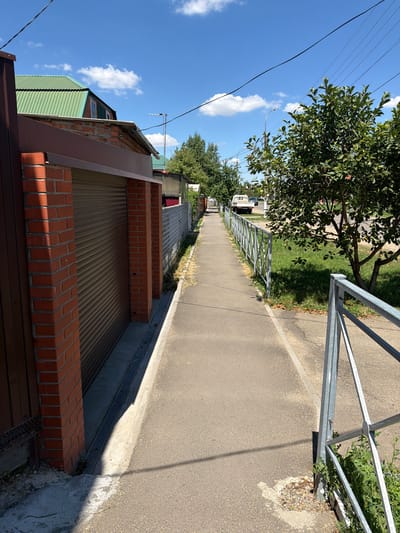 This image depicts a street scene in Timashevsk, Russia, on a sunny day. The perspective is from a low angle, looking down a paved walkway lined with a metal fence on the right and a brick wall with a corrugated metal gate on the left. Beyond the fence, grass and trees are visible, and further in the distance, several houses and parked vehicles can be seen.
The time of day appears to be late morning or early afternoon, judging by the bright sunlight and the shadows cast. The sky is blue with a few fluffy white clouds, suggesting fair weather. The overall atmosphere is peaceful and ordinary, capturing a typical residential street.
Notable details include the construction of the houses, with corrugated metal roofs and brick accents. The metal fence is simple and functional, and the brick wall adds a rustic touch to the scene. There is no visible text in the image. The focus is on the architecture and the urban landscape, with natural elements like trees and grass interspersed. The scene does not appear to capture any specific event or activity; it seems to be a static snapshot of a moment in time.