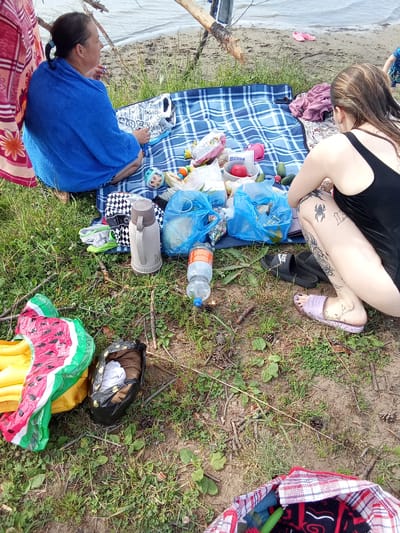 A medium shot captures a group of people having a picnic by the water. In the foreground, a woman in a black swimsuit is sitting on a blue and white checkered blanket, her legs crossed. She has a spider tattoo on her right thigh and other tattoos on her left leg. To her left, another woman is seated with her back to the camera, wrapped in a blue towel. Scattered on the blanket and the grassy ground are various picnic items, including blue plastic bags, a thermos, a bottle of water, a red tomato, cucumbers, an inflatable watermelon, and a patterned bag. In the background, the calm water of a lake or river is visible, with a sandy shore and sparse vegetation. Pink flip-flops are visible on the sand in the distance. The scene is set outdoors on a sunny day, with a general atmosphere of leisure and relaxation.
