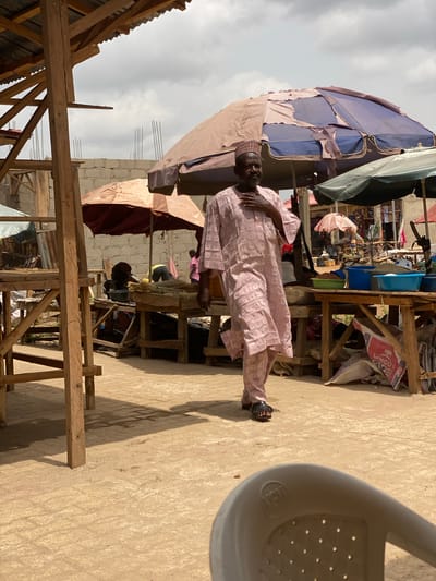 The image shows an outdoor scene in what appears to be a marketplace. An elderly man in a pink patterned robe and cap stands facing the camera. A dilapidated market stall with blue and beige tarps provides shade. Other stalls and merchandise are visible in the background. Wooden structures, possibly parts of stalls or buildings under construction, are scattered around. The ground is a beige color, possibly concrete or packed dirt. A portion of a white plastic chair is visible in the upper right corner. There are people and merchandise within the market, with a building made of cinder blocks and rebar visible in the background. The location is the city of Kafanchan B, Nigeria.