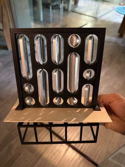 A hand holds a dark-framed, roughly square object containing twelve silver, elongated objects and four smaller, round silver objects arrayed in a grid pattern against a light beige background piece. A dark, multi-tiered rectangular frame extends from the back. The backdrop includes a light wood floor and a stairway descending into a dimly lit area. The location and city of Cholula, Mexico are not deducible from this image alone.