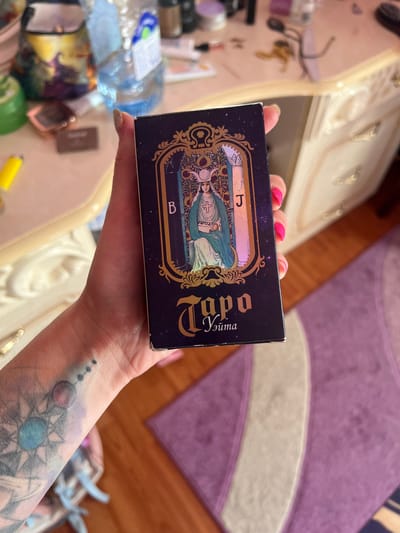 A hand with pink fingernails holds a tarot card box with a dark blue, star-speckled background. The box features an ornate, golden border around an illustration of a robed figure with a halo, holding a staff. The text on the box appears to be in Cyrillic script, with the words "Торо" and "Элита" visible. The background is blurry, showing a wooden floor and a purple rug. Other objects are also out of focus, including a plastic bottle of water and what appear to be cosmetic items. The scene suggests an indoor setting, possibly a home, given the presence of personal items and furnishings. The lighting is soft, indicating it could be daytime or evening. The hand holding the box has a tattoo on the forearm, partially visible. The overall impression is a close-up shot of a hand displaying a tarot card box in a domestic environment.