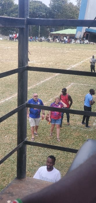 The image captures an outdoor sports ground in Nakuru, Kenya, viewed from an elevated position partially obscured by a dark, metallic structural framework.

In the immediate foreground, a dark-skinned male in a white t-shirt is visible from the chest up, looking upwards. He is positioned above a brown, textured surface.

The midground features a sports field with dry green and brown grass, marked by white lines. On the field, five individuals are present. A light-skinned male with a beard and sunglasses wears a blue jersey and light shorts, walking towards the left while holding a brown bottle. Next to him, a light-skinned female with blonde hair, dressed in a multi-colored t-shirt and denim shorts, also walks leftward, holding an orange object and touching her left ear. Behind them, a dark-skinned male in a red tank top and dark pants stands, facing left. Further to the right, a dark-skinned male in a light blue t-shirt and dark pants stands, oriented left. A fifth dark-skinned male, partially visible on the far right, holds a white spherical object.

The background shows a distant crowd of people dispersed across the far side of the field, some seated. Behind the crowd, a building with a green corrugated roof and an adjacent water tank is visible, set against a backdrop of dense green trees under an overcast sky.