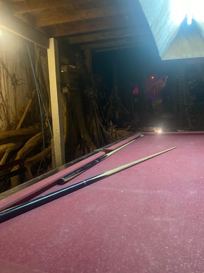 A tilted shot focuses on a red-felted pool table and two pool cues resting on its edge. To the left of the pool table, an obstructed and dark space reveals structural wood and beams, along with a glimpse of red lights in the distance. In the background, an indeterminate area appears cluttered and disorganized. The location is identified as Baan Don Sai, Thailand.