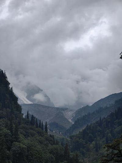 A wide shot captures a vast mountain landscape under a heavy, overcast sky. The foreground is dominated by a dense forest of dark green coniferous trees covering the slopes of a mountain. These trees vary in height and density, with some taller, spire-like evergreens emerging from the lower canopy.
Beyond the forest, the midground reveals rugged mountain terrain. A prominent feature is a light-colored, possibly snow-covered or rocky, slope that descends from a higher peak, appearing like a dried-up waterfall or a scree slope. This area is partially obscured by swirling gray clouds, adding to the atmospheric and dramatic mood of the scene. Further back, additional mountain ranges fade into the misty distance, their contours softened by the clouds.
The sky is filled with thick, textured clouds, predominantly in shades of gray and white, with some areas appearing brighter where light filters through. There are very small, dark specks scattered across the sky, which could be distant birds or insects.
The image is taken from a vantage point looking upwards and across the valley. There are no visible people, structures, or specific landmarks that would immediately identify the location as Burwa, India, though the overall mountainous and forested environment is consistent with some regions of India. The scene is serene and somewhat imposing due to the scale of the mountains and the dramatic weather.