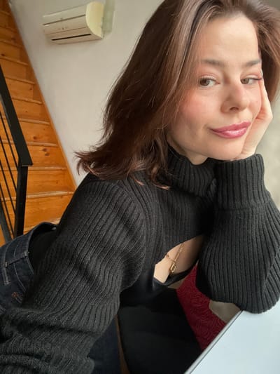 The image depicts a female subject indoors in Roubaix, France. The subject is an adult woman with long, wavy brown hair, light skin, and is wearing makeup, including pink glossy lipstick and defined eyebrows. She has a subtle smile and is looking directly at the camera. Her right hand is placed against her jawline, supporting her head.

She is dressed in a black, ribbed knit, long-sleeved turtleneck sweater that features a significant cut-out across the upper torso and chest. A black garment, possibly a bra or crop top, is visible beneath the sweater's cut-out. A thin gold-colored chain necklace with an irregular-shaped pendant is worn around her neck, visible through the sweater's opening. She is also wearing dark-wash denim jeans.

The background consists of a light-colored wall. To the left, a wooden staircase with a dark metal railing is visible. Mounted on the wall above the staircase is an off-white, wall-mounted air conditioning unit. In the lower right foreground, a white or light grey table surface is partially visible. A cushion with a red and cream wave-like pattern is partially seen behind the subject's left side. The scene is illuminated by ambient indoor lighting.