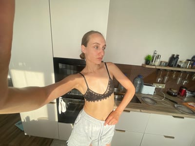 The image shows a woman in a kitchen, standing in front of white cabinets and a black oven. She is wearing a black lace bra and grey sweatpants. Her hair is pulled back in a bun, and she is wearing long earrings. The kitchen counter is cluttered with various items, including bottles, glasses, and a toaster. The lighting suggests it is daytime, with sunlight streaming in from the left. The woman is holding her left arm up and out of frame, as if taking a selfie. The scene is not located in Podshivalovo, Russia.