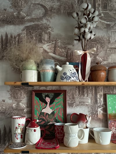 The image depicts a shelf in an interior setting, likely a home, adorned with decorative items and mugs. The background features wallpaper with a sepia-toned landscape of classical architecture and gardens, evoking an old-world aesthetic.
On the top shelf, several objects are arranged: a bunch of dried cotton plant stems in a red vase tied with a pale ribbon, a white teapot with blue floral patterns, a brown acorn-shaped ceramic container, and a ribbed white ceramic container. To the left, there are three stacked mugs, two of which are light blue and pink, and a white mug holding a fluffy, light-colored organic material.
The lower shelf displays an assortment of mugs and a framed artwork. The artwork is a portrait of a woman with dark hair and green foliage, in a dark, richly patterned dress. To the left of the artwork are two festive mugs: one is a tall, white cylindrical mug with a Christmas-themed design of a cat in socks and a candy cane border, and another is a white mug shaped like a Santa Claus gnome. A small round coaster with a mosaic pattern is placed in front of the Christmas mugs. In the center and right of the lower shelf are several more mugs: a red mug with a white rabbit figurine, a white mug designed to resemble a ball of yarn with Russian text that translates to "Morning Tea Love", a plain white mug, and a white mug with a subtle ribbed pattern. A small crocheted pink and red mat is placed near the Santa mug.
The lighting suggests an indoor environment, possibly with natural light coming from a window, casting soft shadows on the shelf. There are no people visible in the image, and no specific actions are depicted beyond the arrangement of objects. The overall impression is one of a curated display of decorative and functional items.
This image does not contain any specific landmarks or contextual information that would allow for its identification or location within the city of Ulyanovsk, Russia. The background wallpaper depicts a generic classical landscape, and the objects themselves are not indicative of any particular geographical origin.