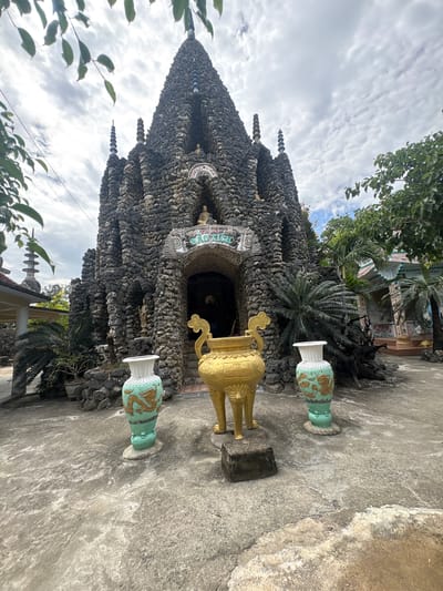 The image captures a unique, intricately constructed grotto-like temple or shrine made predominantly from dark, irregular stones, pebbles, or shells, set in Cam Ranh, Vietnam. The structure rises to a conical or tiered peak, adorned with multiple spire-like elements crafted from stacked circular components. An arched entrance, possibly the main doorway, features lighter-colored shell mosaics, with partially visible stylized text that appears to spell out "THICH CA" or similar, suggesting a Buddhist context.
In the foreground, two ornate decorative urns are placed on a concrete ground. To the left, a large, golden urn stands on stylized legs with an elaborate handle. To its right and further back, another urn is visible, light green and white with gold dragon motifs, standing on a pedestal. Lush green tropical foliage, including palm trees and leafy branches, frames the structure and rises behind it, hinting at a natural, possibly garden setting. The sky above is partly cloudy, indicating daytime, likely under bright, diffused light. No specific activity or people are depicted; the scene is static, showcasing the unique architecture and its surrounding environment.