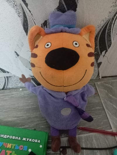 In Novodesyatnikovo, Russia, a plush toy cat character, wearing a purple hat, lavender hoodie, and brown shoes, stands upright on a wooden floor against a textured wallpapered wall. The cat has a large orange head with black ears, big black eyes with white pupils, a black nose, and a smile. A small purple tie with a striped pattern hangs from its neck. To the left of the toy, a green book with Russian text, including "УЧИТЬСЯ" (to learn), is partially visible. A red pen lies on the floor to the right of the toy, with a black cord and a small turquoise object also present on the floor. The lighting in the scene is moderate, casting soft shadows.