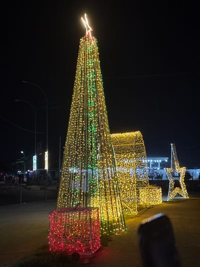 A brightly lit Christmas tree constructed of light strands dominates the night scene. It has a star on top and is decorated with yellow, green, and red lights. Behind the tree are other Christmas decorations, including a star shape and a cylindrical structure, also adorned with lights. The background is dark, with faint outlines of buildings and streetlights. Some people are visible in the distance, adding to the festive atmosphere.