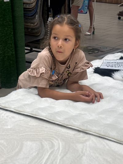 A young girl with braided brown hair and blue hair ties lies on a white rug in a store. She is wearing a light pink t-shirt with ruffled sleeves and a leopard print design on the front. Her arms are resting on the rug, and her hands are clasped together. In the background, there are rolls of green carpet and displays of other rugs. A sign with Russian text is visible on a black and white rug to the right of the girl. The floor appears to be tiled. The setting is consistent with a retail environment selling flooring or home goods.