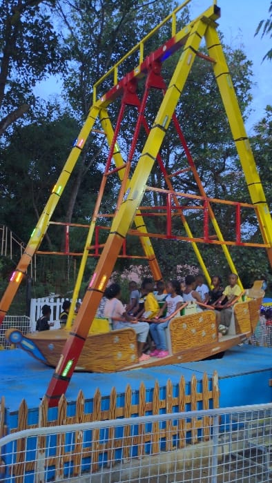 The image depicts an amusement park ride, identified as a "pirate ship" type, situated in the city of Moshi, Tanzania. The scene is captured during twilight or early evening, evidenced by the deep blue sky and the illumination of lights on the ride structure.
The central subject is a large, brightly colored pendulum ride. Its support structure consists of two prominent yellow A-frames connected by a red horizontal crossbar and pivot point. Numerous rectangular LED lights, exhibiting white, green, and yellow hues, are integrated along the yellow support beams and the orange swing arms of the ride. A brown, wood-textured boat-shaped gondola, with elevated bow and stern, is suspended from the red pivot.
Inside the gondola, approximately 8-10 children of varying ages (estimated between 5-12 years old) are seated in rows, facing inwards towards the center of the boat. They are wearing casual clothing. The gondola is observed in a slightly elevated position.
The ride is positioned over a blue ground surface, possibly designed to simulate water, which is enclosed by a decorative, light brown wooden-style picket fence. In the immediate foreground, a grey metal mesh fence is visible, separating the viewer from the ride area. The background features dense green foliage from trees, silhouetted against the dark sky. Faint figures are discernible near a white picket fence in the distant left background.