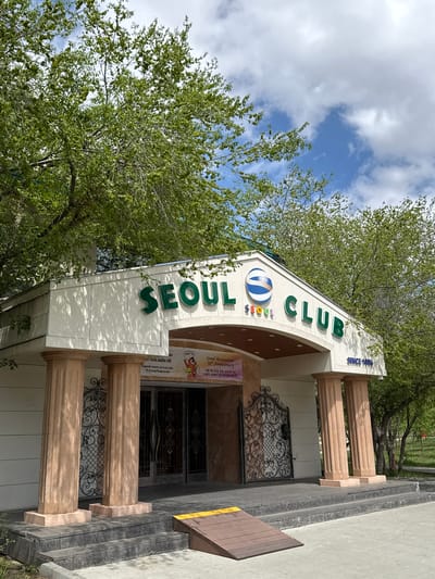 The image shows the entrance to Seoul Club, a restaurant in Ulaanbaatar, Mongolia. The entrance features a white awning with "SEOUL CLUB" in green lettering and a logo. Underneath the awning, a banner announces the restaurant's 30th anniversary in both English and Mongolian. The entrance is framed by tan-colored columns and ornate wrought-iron gates. The sky is visible in the background, partly obscured by trees.