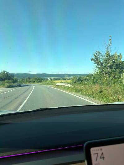 The image, taken from the perspective of a vehicle's dashboard, shows a clear blue sky above a rural landscape. A two-lane asphalt road curves through rolling hills covered in green vegetation and trees. A white guardrail runs along the right side of the road. In the distance, a mountain range is visible. A speed display on the dashboard shows "74 km/h." The scene is bright and sunny, suggesting daytime. The location is Rish, Bulgaria.