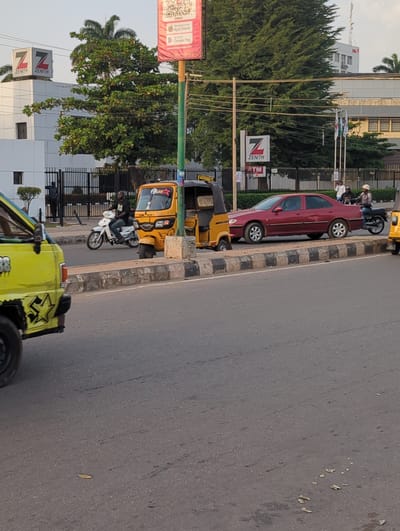 The image depicts a street scene in what is likely the city of Shaba, Nigeria, given the Zenith Bank signage in the background, a prominent bank in Nigeria. Multiple vehicles are visible, including a red sedan parked alongside the road, a yellow auto rickshaw (keke) and a green vehicle. People can be seen on a motorcycle and other unidentified people are standing on the sidewalk nearby. The street appears paved, and the environment suggests an urban setting with trees and buildings visible in the background.