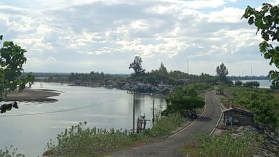 The image shows a view of a coastal area with a river or bay in Baroh Lancok, Indonesia. In the foreground, a paved road curves through lush green vegetation. On the right side of the road, there is a small, rustic wooden shack with a rusted metal roof and blue paneling on its side, situated on a rocky embankment. Further down the road, more trees and vegetation are visible.

The mid-ground features a body of water where numerous fishing boats are docked. These boats are of varying sizes and colors, predominantly blue and green. Along the water's edge, there are some buildings that appear to be part of a settlement or fishing village. Telephone poles and overhead wires are also present, indicating infrastructure.

The background depicts a distant treeline, with some taller trees and what appear to be communication towers scattered across the landscape. In the far distance, a faint mountain range can be seen under a partly cloudy sky. The water's surface reflects the sky, creating a shimmering effect. The overall scene suggests a working fishing community situated in a tropical coastal environment.