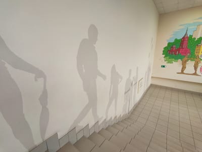 This media file depicts a hallway with a staircase and a mural. The hallway walls are adorned with silhouette-like stencils of people walking, suggesting movement or a procession. One of the silhouettes appears to be holding an umbrella. The stencils are in various shades of gray, blending into the white wall.

On the far wall, a colorful mural is visible. It features a stylized depiction of a city with a prominent red-domed church, green foliage, and tall buildings. Two figures are painted in the foreground of the mural, facing each other. An exit sign with an arrow pointing to the right is also present near the mural.

The staircase is tiled with light-colored square tiles, with a lighter colored border along the edge of each step. The scene appears to be indoors, possibly in a public building such as a school or community center, given the presence of the mural and the general decor. The lighting suggests it is daytime, but there are no explicit cues about the weather or time of day beyond the indoor setting. No distinct emotions are conveyed by the static imagery. The location context provided is Kurgan, Russia.