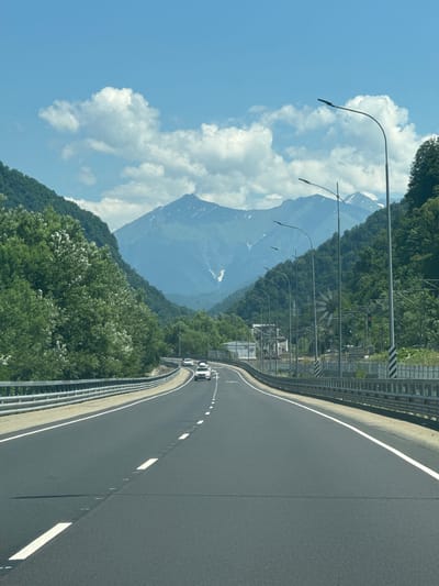 The image captures a view from a vehicle traveling on a paved highway that winds through a mountainous, forested region. The highway has clear white lane markings and a metal guardrail along its edge. On either side of the road, lush green trees cover the slopes of the mountains, with some patches of white flowers visible on the left side. In the distance, majestic mountains rise, some with snow-capped peaks, under a bright blue sky with scattered white clouds. Several cars are visible on the highway ahead, and streetlights line the road. The overall scene suggests a scenic drive through a natural landscape, likely in or near Chvizhepse, Russia, given the mountainous terrain and vegetation.