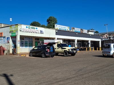 The image shows a street scene in Gitaru, Kenya (latitude: -1.231, longitude: 36.668). There is a pharmacy on the left side of the image, with a sign that says "The Master Pharmacy 24/7". Next to the pharmacy is a larger building with a sign that says "CC Cafe & Bakery" and another sign that reads "CC Liquor". There are two cars parked in front of the buildings, a black car with its trunk open and a beige pickup truck. A man wearing a blue shirt is loading items into the black car. The buildings are situated on a paved street with a brick-like pattern. The sky is clear blue. There are trees in the background. There is a red fire hydrant near the corner of the pharmacy building. There are yellow poles in the street. There is a pedestrian walking on the sidewalk in front of the pharmacy. The street appears to be fairly quiet, with only a few people visible.