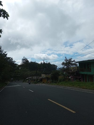 The image depicts a road scene in Phoxay, Laos, under a sky with significant cloud cover and patches of blue. A dark asphalt road, featuring a solid yellow centerline in the foreground and broken white lines further ahead, curves to the left into the distance. White dashed markings are also present on the far left side of the road. Several motorcycles with riders are visible on the road: one in the mid-ground approaching the right side of the frame, and at least two more further down the curving road.

On the right side of the road, adjacent to a grassy bank, multiple structures are present. The most prominent is a two-story green building with a red roof and a wooden balcony on the second floor. To its left, a partial view of a lighter-colored, possibly pink, building is visible. Further left, a yellow single-story building features a covered outdoor seating area and visible but illegible signage, consistent with a commercial establishment. Utility poles supporting multiple electrical wires stretch across the upper right quadrant of the image.

The left side of the road is bordered by dense green foliage and trees that extend into a forested hillside. In the background, beyond the treeline, communication towers are discernible atop a distant elevated landform. The overall environment suggests a semi-rural or mountainous setting.