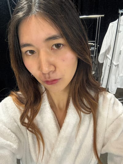 A young woman is captured in a close-up, chest-up shot, looking directly into the camera. She has long, dark brown hair with lighter auburn highlights, which falls over her shoulders and partially frames her face. Her head is tilted slightly to her left, creating a candid pose. She is wearing a white, textured bathrobe, suggesting a relaxed or post-bath setting. Her complexion is fair, with a natural, rosy flush on her cheeks, and her lips are colored with a pinkish-red tint. Her eyes are dark brown and meet the viewer's gaze with a calm, neutral expression.

In the background, out of focus, there is a metallic rack, possibly a towel or clothes rack, draped with several white towels or linens. The setting appears to be indoors, likely a bathroom, hotel room, or domestic environment, given the presence of the bathrobe and towels. The lighting is soft and even, suggesting indoor artificial light or diffuse natural light. There is no visible text in the image. The overall impression is one of relaxed intimacy.