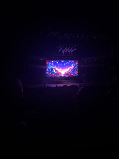The image shows a large, rectangular LED screen displaying an abstract, colorful image in a dark environment. The screen is supported by a metal framework, suggesting it is part of a stage setup. Various lighting fixtures and rigging elements are visible around the screen. The overall setting appears to be an indoor concert or performance venue in Cholula, Mexico. The limited visibility suggests a dark, low-light condition, typical of such events. In the foreground is a blurred silhouette of a head of a person in the audience.