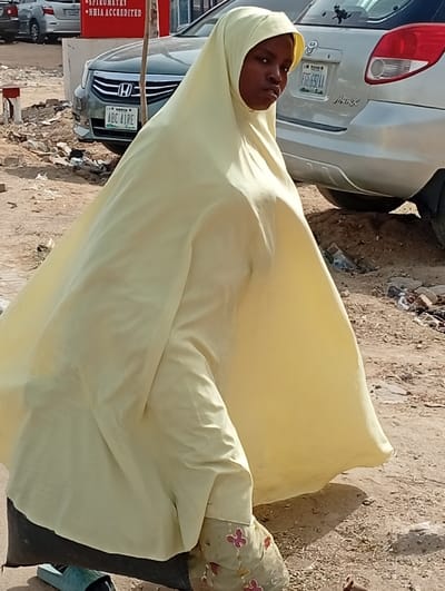 A single individual, appearing female, is positioned centrally in an outdoor, unpaved urban environment in Fagge D2, Nigeria. The individual wears a pale yellow, full-body garment covering the head, torso, and limbs. Their dark-skinned face is visible, with eyes directed towards the viewer. Light-colored footwear adorned with red, butterfly-like embroidery is visible on the feet, beneath the garment's hem. A dark-colored sandal or slipper rests on the dusty, light brown ground to the left of the individual's left foot. The ground exhibits scattered debris. In the background, multiple vehicles are parked. A silver Honda sedan, bearing license plate "KANO ABC 41RE", is partially visible behind the individual to the left. Further right and behind, a silver SUV is parked. A red rectangular sign with white text, including "SPIROMETRY" and "NHIA ACCREDITED", is partially visible to the upper left. A thin vertical pole is positioned near the red sign. The scene is brightly illuminated by daylight.
