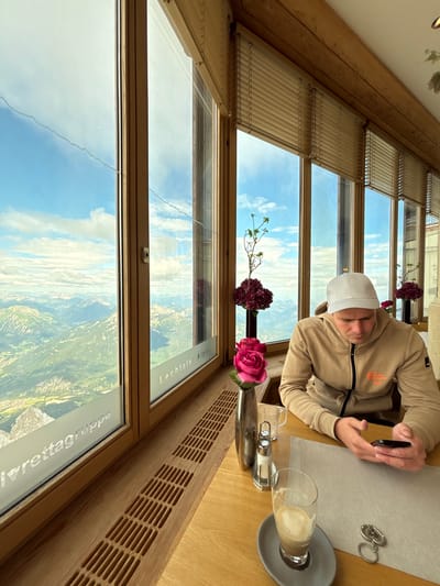A man wearing a light-colored baseball cap and a tan hooded sweatshirt is seated at a wooden table, looking at his smartphone. He is positioned in the foreground, to the right of the frame, with a glass of coffee and a salt and pepper shaker on the table in front of him. Behind the man, a large window offers a panoramic view of a mountain range under a partly cloudy sky. The window is framed by wooden beams and blinds. The text "Lechtaler Alpen" and "lvreittagruppe" are visible on the window. The setting appears to be a restaurant or cafe with large windows, likely at a high elevation in the Austrian Alps near Ehrwald, Austria.