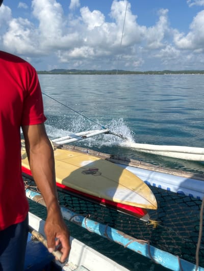 The image captures a view from a moving boat in General Luna, Philippines. The sky is visible with scattered clouds above the ocean. A section of land covered with vegetation is present on the horizon. On the right side of the frame is a portion of the boat structure, including blue-painted wooden poles, netting, and a surfboard secured alongside. Water splashes against the boat's side. In the foreground, a person wearing a red shirt is seen holding onto part of the boat's frame.