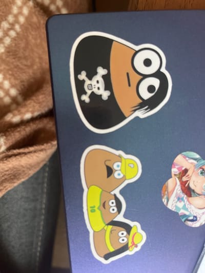 The image displays a close-up, top-down view of stickers on a dark blue laptop. The stickers are cartoonish in style. One sticker features a character with a black background, a skull and crossbones symbol, and a brown face with two large white eyes with black pupils, and a thin black line for a mouth. Another sticker depicts a stack of three characters with brown, bean-shaped heads. The top character wears a yellow hat with a white 'p' on it. The middle character wears a bright yellow shirt with the number '10' visible in black. The bottom character wears a yellow hat with a pink ribbon. To the right of these stickers is a circular sticker of an anime-style character with blue hair and large green eyes, wearing a colorful outfit. The background of the image shows a brown and white patterned fabric and the edge of a wooden surface. There is no indication of the location being in Ivano-Frankivsk, Ukraine, or any other specific city.