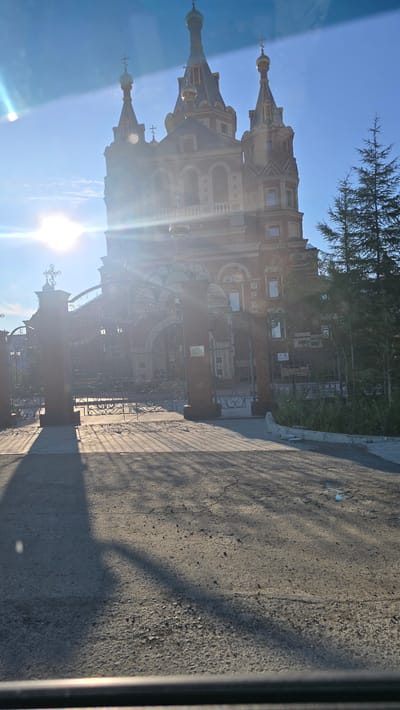 An exterior shot captures a large, red brick building with multiple towers and golden domes, likely a church or cathedral, under a bright blue sky. The sun is shining intensely, creating lens flare and casting long shadows from a decorative wrought-iron gate in the foreground onto the asphalt ground. A coniferous tree is visible to the right of the building. The building features intricate architectural details, including arched windows and ornate brickwork. The overall scene suggests a sunny day in a city, possibly during the early morning or late afternoon given the long shadows. The location is identified as Udachny, Russia.
