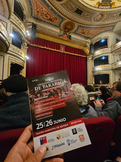 The image shows a person's hand holding a program for an orchestral concert in the Teatro Municipal de Iquique, Chile. The theater's ornate interior, with gilded balconies and red velvet seating, is visible in the background. The program details the concert by the Orquesta Regional de Tarapacá, featuring works by Camilo Roca, Richard Strauss, and Antonio Vivaldi. The concert is scheduled for June 25th and 26th, 2026, at 8:00 PM. Several audience members are seated in the foreground, some looking at their phones.
