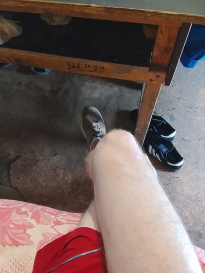 The media file shows a person's leg and foot, wearing a gray sneaker with white laces. The person is sitting on a pink and white floral-patterned cushion, with red shorts visible. The background features a wooden table with a dark interior and some items within. On the table's wooden surface, there is some text that appears to read "3AA 26.03.08 12.22".
Next to the table, on the ground, there are a pair of black Adidas sneakers with white stripes and a blue emblem. The floor is concrete and appears somewhat worn.
The scene is indoors, possibly in a casual or rustic setting. The lighting suggests it might be daytime. The image does not convey any specific emotion. The location context provided is Magnitogorsk, Russia.