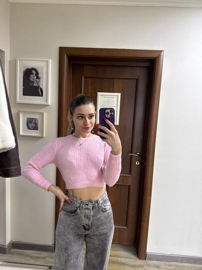 An individual is depicted taking a mirror selfie indoors in Reutov, Russia. The subject is a young adult female with dark hair pulled back, light skin, and a slight smile. She wears a pink, cropped, ribbed long-sleeve sweater, acid-wash grey high-waisted jeans, and has a visible tattoo below the hem of her sweater on her left side. Her left hand rests on her hip, displaying red nail polish and a silver-colored ring on her ring finger. Her right hand holds a dark-colored smartphone, equipped with a purple case, pointed towards the mirror. She also wears earrings and a delicate necklace.
The background consists of a light-colored, possibly cream, wall. To the left, a dark brown jacket with a white/cream fur-lined collar or interior hangs on a rack. Above it, two framed black and white photographs are mounted vertically: the upper one depicts a woman with dark hair smoking, and the lower shows a woman's face. Directly behind the subject, a dark brown wooden door with a brass-colored handle and lockplate is visible. A white-framed document or certificate, featuring Cyrillic text (e.g., "АТТЕСТАТ" at the top), is affixed to the door. The floor appears to be a light-colored wood or laminate. The scene is illuminated by ambient indoor lighting.