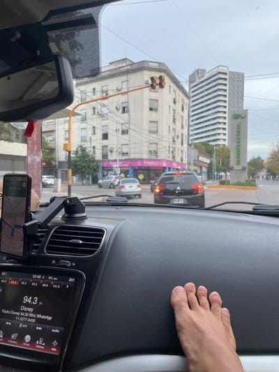 The image shows a view from inside a car, likely in Avellaneda, Argentina, as the radio is tuned to Radio Disney and WhatsApp is indicated on the radio display. A navigation app showing a route on Av. Manuel is visible. The building with the sign "YA ESTAS EN CASA" (You Are Home) is seen through the windshield. The sky is overcast. A person's foot is resting on the dashboard.