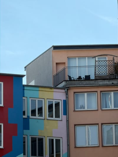 This image shows a close-up, Dutch angle view of the exterior of a multi-story building, with sections of the sky visible. The building features several floors with different colored facades and multiple windows. The top section has a light peach-colored facade with a dark metal railing and a decorative metal screen on a balcony. Below this, a section of the building is a pale pinkish-beige color. Further down, the facade is painted with large, colorful geometric blocks of yellow, light blue, and a darker teal, with rows of white-framed windows interspersed. The lowest visible section is painted in bold red and blue blocks.
Several windows are visible, mostly rectangular and white-framed. Some windows appear to have blinds or curtains. On the balcony of the upper floor, black chairs are visible, suggesting it's a functional outdoor space. The sky is a clear, pale blue, indicating daytime with fair weather. The overall impression is of a modern or renovated apartment building, possibly with a vibrant and artistic design. The angle of the photograph, looking upwards and tilted, adds a dynamic and slightly abstract quality to the scene. There is no visible text in the image. The context suggests this is a building in Frankfurt, Germany.