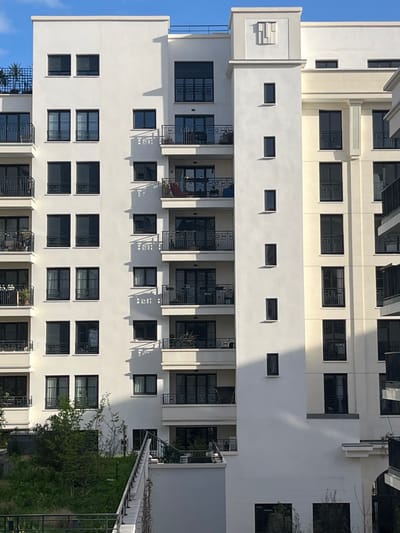 The image depicts a multi-story residential building complex located in Saint-Ouen-sur-Seine, France, under clear blue skies. The primary structure features a predominantly white facade with dark-framed windows arranged in a grid pattern. Multiple balconies, equipped with black metal railings, are present on most floors; some contain potted plants, and one balcony on a middle floor includes red and blue outdoor chairs.
A prominent vertical architectural element, also white, extends through the height of the building, featuring five small, rectangular, vertically aligned windows and capped with a lighter grey decorative frieze-like structure at the roofline. Adjacent to this, a section of the complex exhibits a cream-colored facade with vertical pilasters and similar dark-framed windows and balconies. The scene is illuminated by direct sunlight, casting sharp, diagonal shadows from balconies, window frames, and architectural protrusions onto the building surfaces.
At the ground level, green foliage, shrubs, and an upward-sloping grassy area are visible. A set of outdoor stairs with a grey concrete base and black metal railings ascends diagonally from the lower left towards the building. No individuals or active interactions are visible.