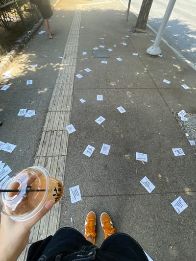 The image depicts a sidewalk in Thessaloniki, Greece, littered with numerous small white flyers. A person's legs, wearing orange sneakers and dark pants, are visible in the foreground, holding a clear plastic cup with an orange beverage and a black straw. The cup has text that appears to read "I NEED". The sidewalk has tactile paving along its edge. In the background, a person wearing sandals and dark shorts is walking away from the camera, casting a shadow. Sunlight filters through trees, creating dappled light patterns on the ground. The flyers are scattered across the concrete, some bearing Greek text.