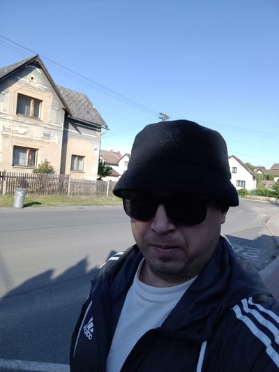 A man wearing a black bucket hat and sunglasses stands on a paved road in Čistá, Czechia. He is wearing a white t-shirt and a black Adidas jacket with white stripes on the sleeves. The man is looking at the camera. Behind him, there is a house with a wooden fence and a trash can. The sky is clear and blue.
