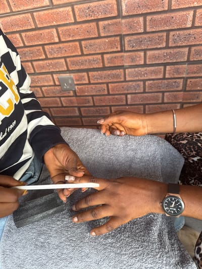 The image shows a close-up view of two people's hands, with one person filing the nails of the other. The background features a brick wall pattern and a grey textured surface, possibly a towel or blanket. One person is wearing a watch with a black strap and a rose gold casing. The other person is wearing a striped shirt with black and white bands, and a yellow and white emblem with text on it. The activity taking place is a nail filing session, likely at a salon or in a home setting. The overall lighting suggests daytime. The location context is Ajara, Nigeria. There is visible text on the emblem of the striped shirt, but it is partially obscured and difficult to read clearly. The hands suggest a person of color.