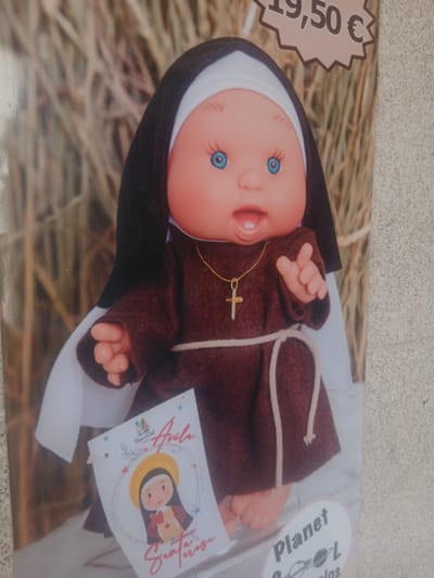 A doll dressed as a nun is shown in a photograph. The doll has blue eyes and a smiling mouth with visible teeth. It wears a brown habit with a white veil and a gold cross necklace. A beige rope belt is tied around its waist. In the lower left corner, a small card displays a cartoon image of a nun with a halo and the text "Aula Santa Teresa" and "Planet L". The background appears to be made of dried grass or straw. A price tag in the upper right corner reads "19,50 €". The city of Ávila, Spain, is famous for being the birthplace of Saint Teresa of Ávila, the subject of the doll.