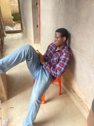 A young, dark-skinned man sits on an orange chair, leaning forward slightly. He wears a red, blue, and white plaid shirt with blue jeans. He has short, dark hair. The background includes a worn, painted wall and ceiling. A window shows an exterior view of green foliage and scattered refuse. The perspective is unusual, as if the image was rotated.