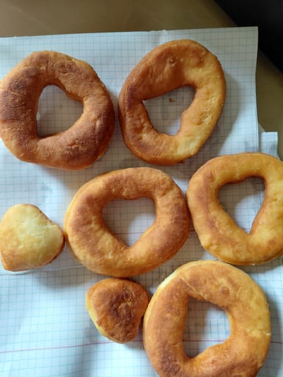 A top-down, eye-level shot captures seven golden-brown, irregularly shaped fried dough pastries laid out on a piece of grid paper. Six of the pastries are donut-shaped with a hole in the center, and two are heart-shaped. The pastries appear to be slightly undercooked in places, with a matte, slightly bubbly surface. The grid paper is white with blue lines and red horizontal lines, suggesting it might be a notebook page. The background is a neutral, light brown surface, possibly a table or desk. There are no people or animals visible in the image. The lighting is bright, creating soft shadows beneath the pastries. The scene is a close-up, focused entirely on the food items and the paper they rest on. This scene is not specific to Oskemen, Kazakhstan or any other city.
