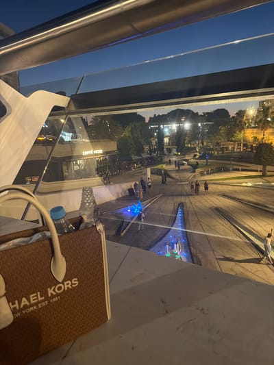 The image shows an outdoor scene at dusk, possibly in Nicosia, Cyprus. In the foreground, a brown patterned bag with "MICHAEL KORS NEW YORK EST. 1981" printed on it rests on a white surface. A blue-capped water bottle and a white cable are also visible.

Beyond the foreground, a wide-angle view reveals a public space with a fountain and many people. The fountain is illuminated with blue lights, and water jets are visible. Several individuals are standing around the fountain and walking through the paved area, suggesting a casual gathering or leisurely activity. The time of day is dusk, indicated by the darkening sky and the artificial lighting in the scene, including lights from buildings and street lamps.

Further in the background, trees and buildings are visible. One building has a sign that reads "CAFFE NERD". The overall atmosphere appears relaxed and social, with people enjoying the evening in a public plaza. The lighting creates a slightly dramatic effect, highlighting the fountain and the architectural elements of the space.