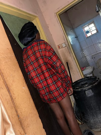 The image depicts an interior scene in Muchiya, Nigeria, focused on an individual standing in a doorway. The person, seen from behind, has dark skin, wears a black satin bonnet, and an oversized red, black, and blue plaid button-up shirt that extends to mid-thigh, leaving the lower legs bare.
To the immediate left, a light brown/tan wall or doorframe is visible, partially obscured by a light gold fabric or curtain. The floor beneath the individual is dark.
To the right, a doorway opens into an adjacent room, likely a bathroom, characterized by light blue and white tiled walls. Within this room, a white toilet with an open seat is visible, along with a white bidet or washbasin above it. A small, barred window is present high on the far wall of this inner room, with a dark item suspended near its bars.
In the foreground, to the individual's right, a large black plastic drum with an open top rests on the floor. A dark rod, possibly a broom handle, leans vertically against the wall between the person and the drum. Two electrical switches or outlets are mounted on the wall within the doorway frame, surrounded by areas of discoloration.