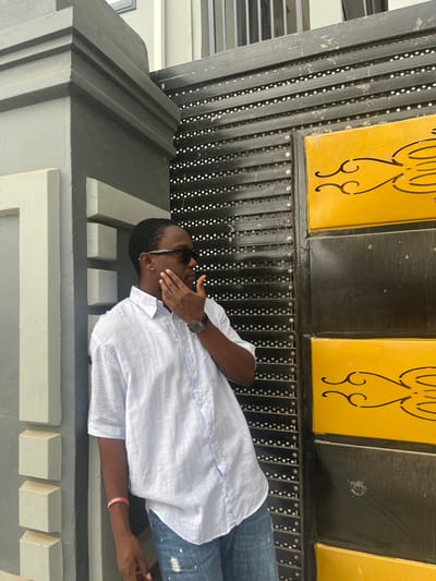 A person wearing a white button-down shirt, jeans, and sunglasses reclines on a decorative, grey and white surface. The individual covers part of their face with their hand. Above the person is a structure of black metal with rows of holes, decorated with yellow panels featuring curved designs. A watch is visible on the person's wrist and a pink wristband on the other.