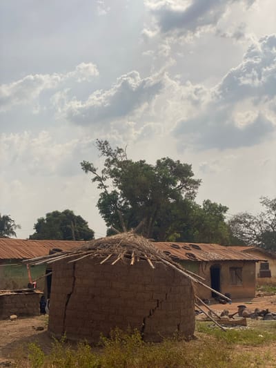 This image depicts an outdoor scene in Nev, Nigeria, under an overcast sky with scattered clouds. In the foreground, dry brown vegetation is visible. The central subject is a circular, single-story structure constructed from mud bricks, exhibiting prominent vertical cracks on its exterior. Its conical roof is partially covered with straw or reeds, supported by visible wooden poles. To the immediate left and slightly behind this structure, a smaller, lower mud-brick wall or partial structure is present, behind which a dark-skinned individual wearing a dark blue garment is partially visible, facing left.

Behind the central circular structure, a rectangular single-story building is visible with walls appearing to be light-colored. This building features a rusted, corrugated reddish-brown metal roof that shows signs of wear, discoloration, and several dark, circular objects (possibly tires) resting on it, along with multiple smaller indentations or holes. An open doorway, appearing dark, is visible on the building's facade, along with a window fitted with wooden shutters to its right. The ground surrounding the structures is dry, dusty earth. In the mid-ground right, a collection of objects including wood scraps and stones is scattered on the ground near the rectangular building's entrance. The background features dense green foliage from several large trees, with one prominent tree rising significantly above the buildings. Additional structures with similar corrugated metal roofs are faintly visible further in the background to the right.