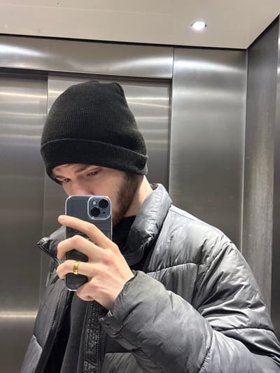 A man wearing a black beanie and a gray puffer jacket takes a selfie. He holds an iPhone in a clear case, revealing dual camera lenses. A gold ring adorns his left ring finger. The background appears to be the interior of an elevator, with stainless steel panels and a glossy white corner. The image is slightly rotated.