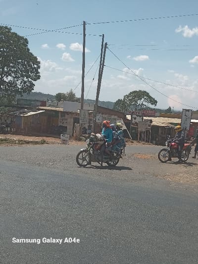 This outdoor image, captured in Kipkarren, Kenya, depicts a daytime scene on a paved road with clear skies. The photograph is oriented vertically, but the content appears to have been shot in landscape.

In the foreground, a motorbike carries multiple passengers, with at least three individuals clearly visible, including the driver and passengers, suggesting shared transport. Further down the road, another motorbike with a single rider is partially visible. Distinct shadows cast by the utility poles and motorbikes indicate sunny weather.

To the left of the road, several wooden utility poles with numerous power lines stretch across the frame, silhouetted against a bright blue sky adorned with white, puffy clouds. The background features green trees and what appears to be elevated terrain or hills.

On the right side of the road, a series of structures, likely small shops or market stalls, are lined with various signs and posters. While most text is unreadable due to distance and resolution, words like "Mpesa" and partial phone numbers (e.g., "07...") are faintly discernible, pointing to local businesses and services.

The scene captures a typical moment of daily life and transportation in a semi-urban or rural Kenyan setting. A "Samsung Galaxy A04e" watermark is present.