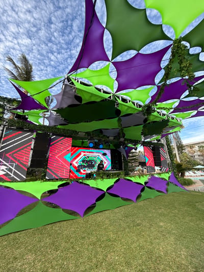 This image depicts an outdoor event setup located in Noumea, France. A stage is situated at the far end of a manicured green lawn, beneath an expansive, geometrically patterned canopy of tensioned fabric. The canopy consists of tessellating lime green and deep purple four-pointed star or diamond-shaped panels, supported by a metal truss system. Green leafy vines are draped over sections of the trusses and hang from the canopy.
On stage, a single individual, appearing to be a male in a dark t-shirt, stands centrally behind a DJ console, actively operating equipment. The stage is flanked by tall speaker stacks and features multiple large LED screens displaying vibrant, abstract geometric patterns in red, black, white, blue, and green. Additional stage lighting fixtures emit blue light. The front of the stage and its upper perimeter are decorated with matching lime green and purple fabric panels and green foliage.
In the background to the right, a multi-story building, consistent with a resort or hotel, is visible. In front of this building, a swimming pool area can be observed, complete with blue water, white lounge chairs, and light-colored umbrellas. Palm trees and other tropical vegetation are present in the immediate vicinity of the background structures. The sky is partially cloudy with scattered white clouds against a blue backdrop, indicating daytime conditions.